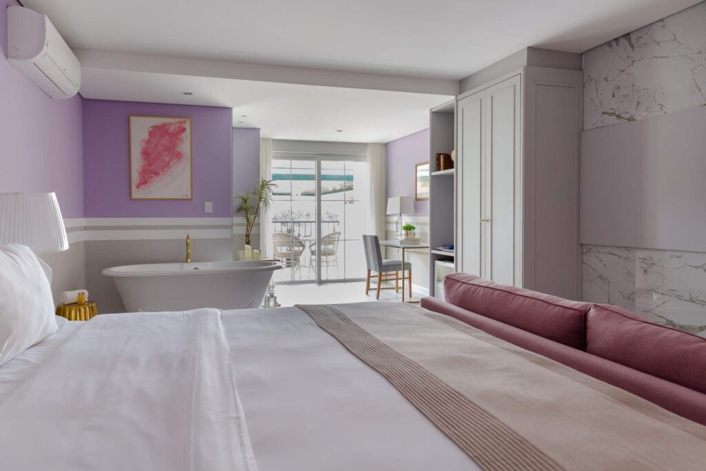 Imagem de um quarto grande com uma banheira de imersão vitoriana vestindo o espaço, o quarto possui tons rosas e lilás