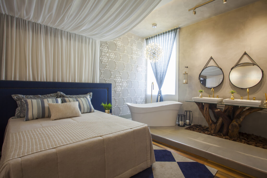 Imagem de um quarto onde aparece uma banheira abaixo da janela ao lado da cama. também possui duas cubas em cima de um tronco de árvore. 