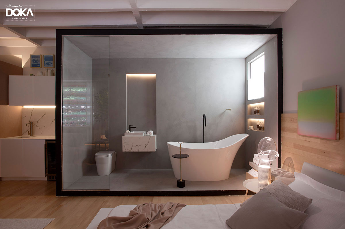 Imagem de uma banheira dentro do quarto, a banheira está dentro de um espaço de vidro, como se fosse um box grande, com uma cuba supensa e um vaso sanitário no mesmo espaço. a imagem mostra uma parte da cama também.