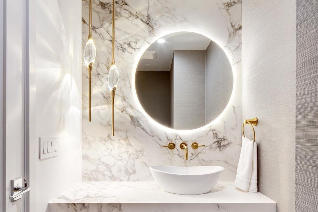 imagem de um lavabo bem iluminado, com metais dourados e uma parede de porcelanato e um espelho redondo com iluminação de led em volta.