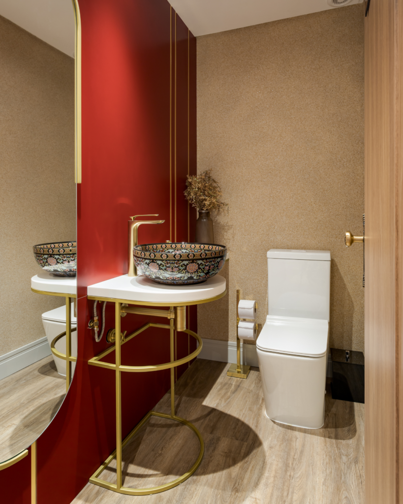 Banheiro com parede vermelha e um espelho na parede. A cuba desenhada está sobre uma bancada com pés dourados e mesa branca. O vaso sanitário é branco e o espaço possui uma papeleira dourada e outros detalhes dourados. 
