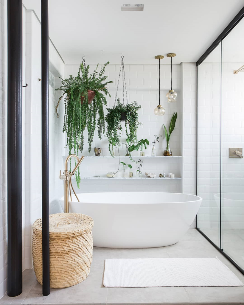 sala de banho branca com banheira de imersão instalada. em cima da banheira existem plantas penduradas e atrás da banheira tem prateleiras com mais plantas. 