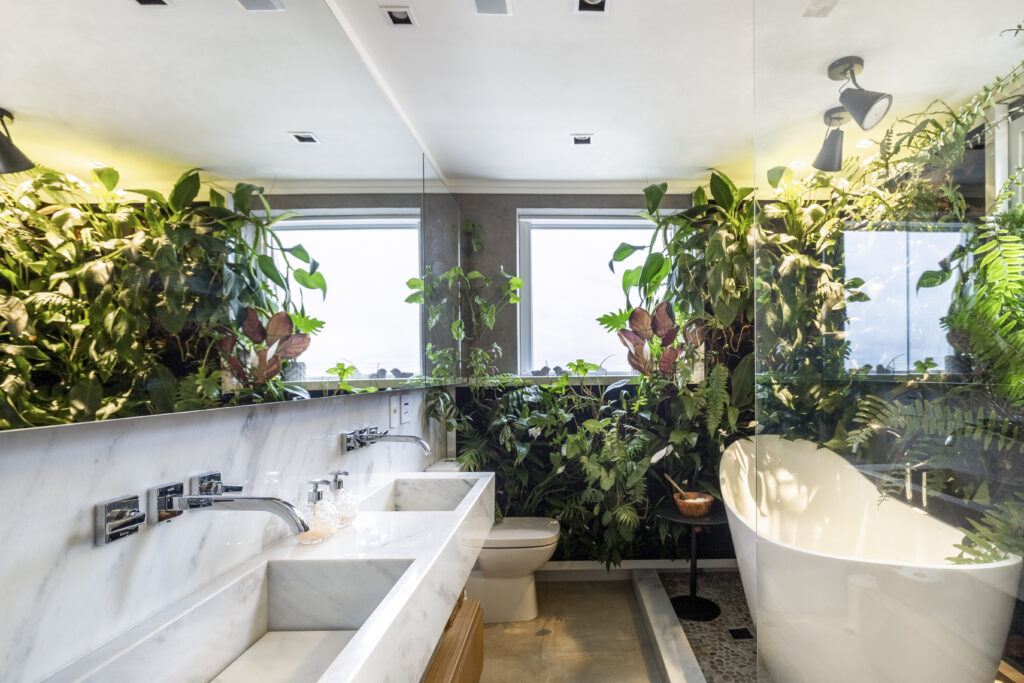 sala de banho com muitas plantas. possui um lavabo duplo feito em mármore, um vaso sanitário e uma banheira branca com muitas plantas verdes em volta 