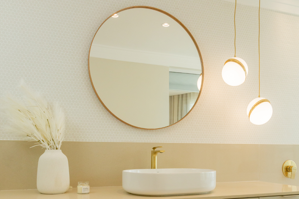 imagem de um lavabo com cores brancas e douradas. uma bancada com uma cuba branca. um espelho redondo e ao lado um lustre pendente dourado. 