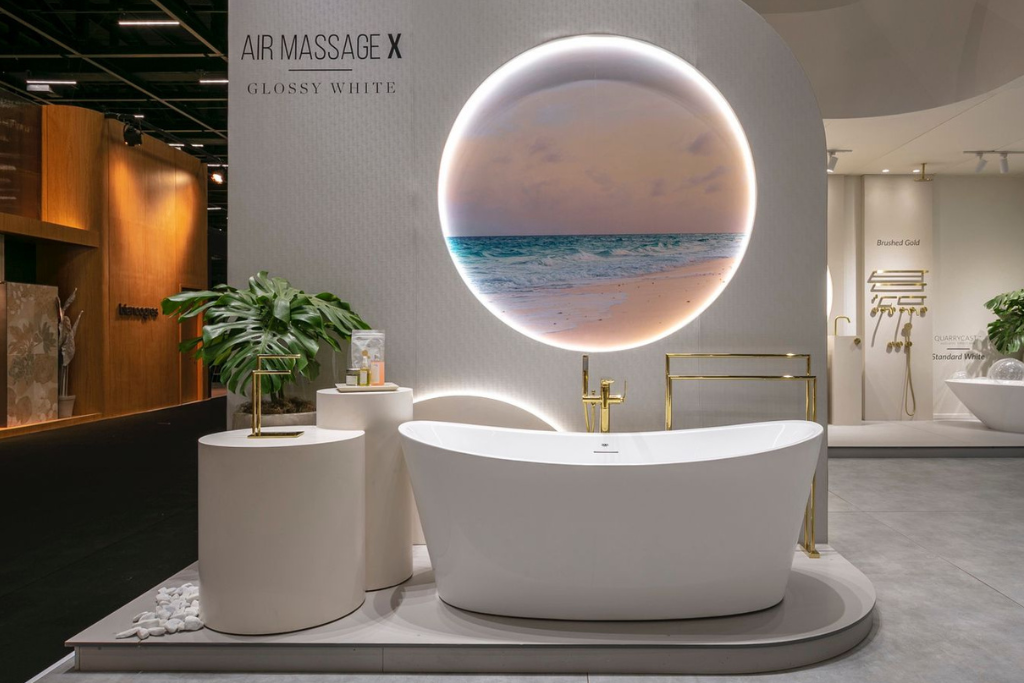 banheira de imersão branca com design moderno e minimalista. Atrás dela tem uma tela redonda com led em volta, a imagem da tela é uma paisagem de praia. Os metais que compõem o ambiente são dourados. 