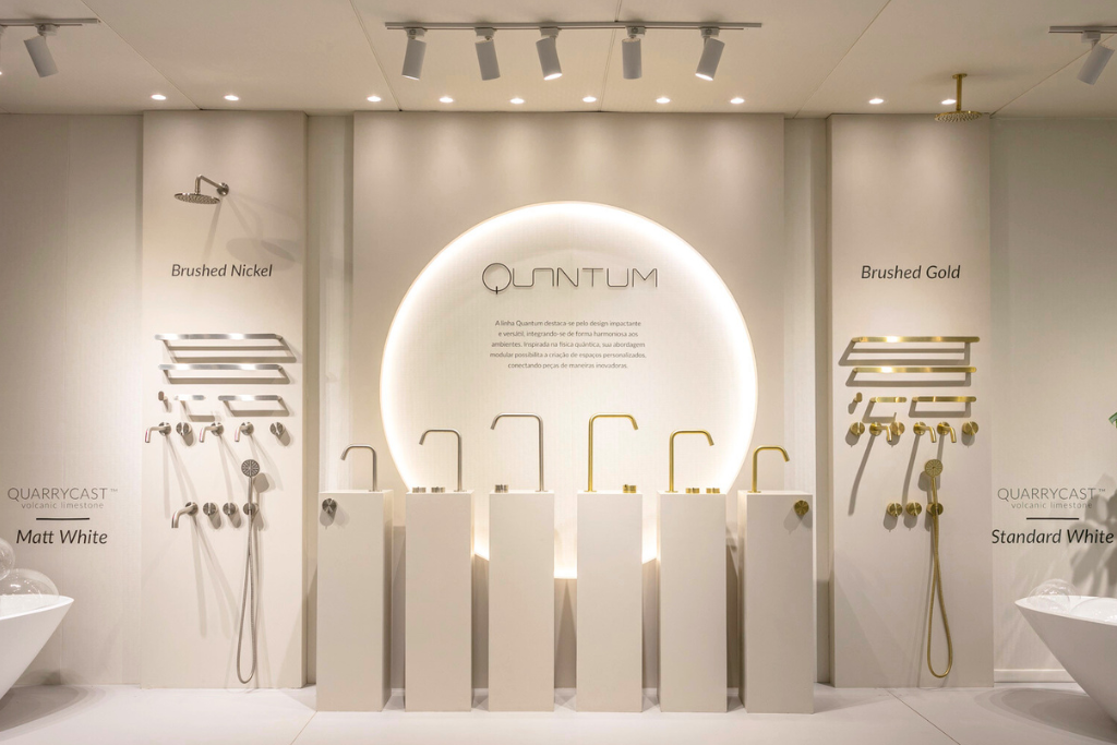 Painel bem iluminado de metais da linha Quantum exposta no estande da Doka, na Revestir 2024; na parede há um circulo e dentro dele o nome da linha e o texto sobre ela. Na frente do circulo temos seis bancadas com os monocomandos em cima de cada um e os volantes também. 