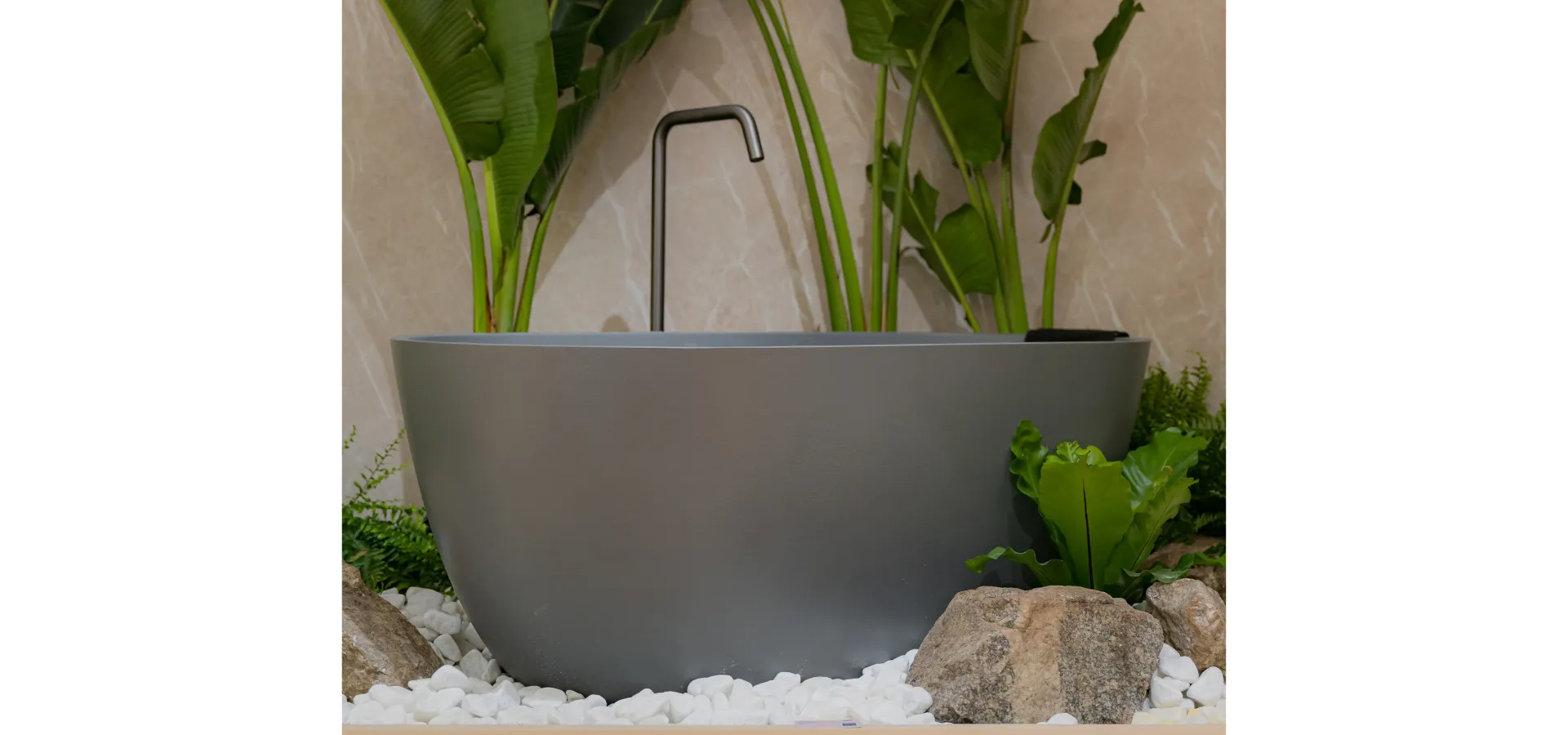 banheira freestanding Texture Doka em acabamento cinza fosco com bica de piso para banheira Quantum Doka em acabamento grafite, cercada por pedras brancas, rochas e plantas tropicais — imagem do artigo: bica ou monocomando para banheira freestanding