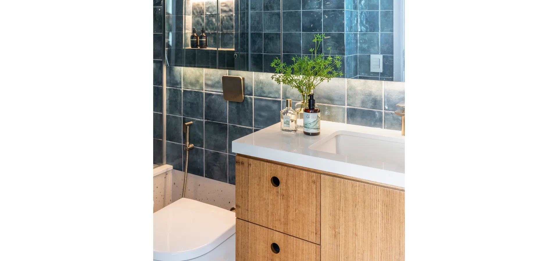 `banheiro com revestimento em azulejo artesanal azul escuro, ducha higiênica Doka em acabamento escovado instalada na parede ao lado de vaso sanitário branco, bancada branca com cuba esculpida, móvel em madeira natural com puxadores redondos e planta verde decorativa`