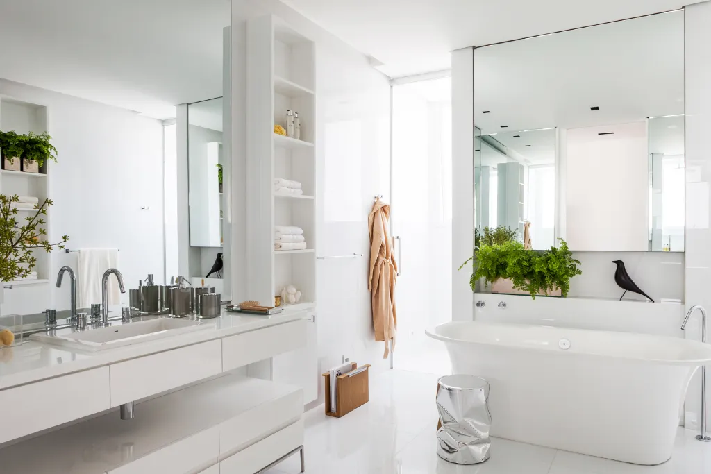 Banheiro moderno e elegante com design minimalista em tons de branco. À direita, há uma banheira de imersão freestanding diante de um grande espelho, com uma planta verde sobre o tampo ao fundo. Ao lado, uma escultura preta em forma de pássaro decora o ambiente. À esquerda, uma bancada branca com pia embutida, metais cromados e acessórios organizados. Prateleiras abertas com toalhas e itens decorativos completam o espaço. A iluminação natural entra por uma ampla janela, realçando o brilho do ambiente.