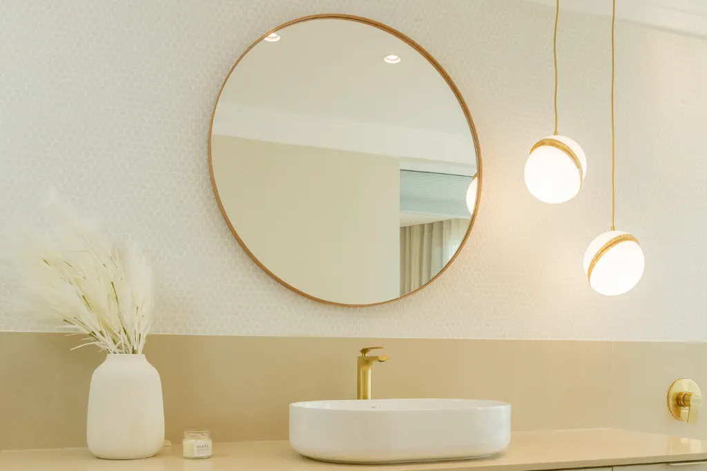 Bancada de banheiro elegante em tons neutros, com cuba branca de apoio e torneira dourada. Acima da bancada, há um espelho redondo com moldura fina de madeira e duas luminárias pendentes com globos brancos e detalhes dourados. À esquerda da cuba, um vaso branco com arranjo de flores secas claras e uma pequena vela decorativa completam a composição, criando uma atmosfera leve e sofisticada.