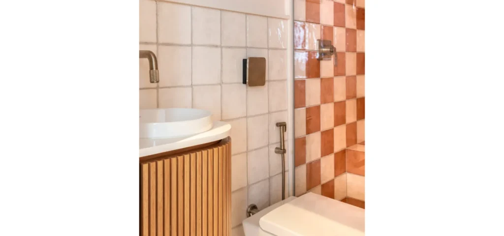 banheiro com revestimento xadrez em tons terrosos, vaso sanitário e ducha higiênica Doka em acabamento escovado instalada na parede, ao lado de cuba de apoio branca sobre móvel ripado em madeira e torneira escovada