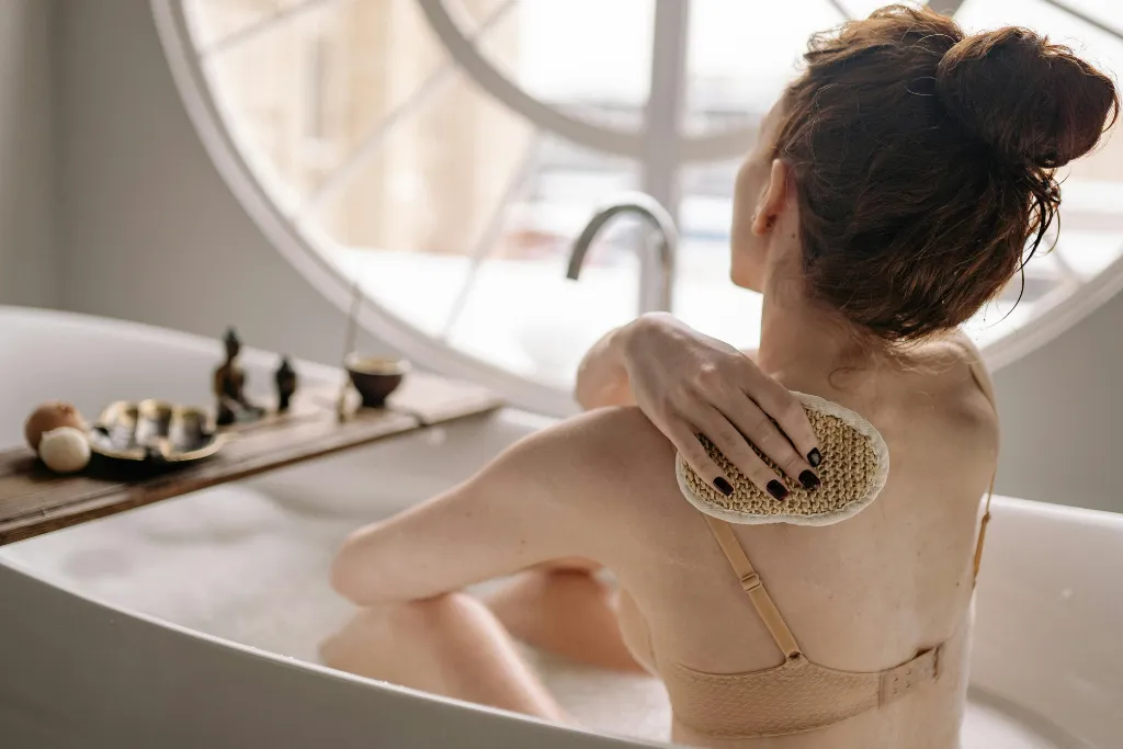 Mulher relaxando em uma banheira, esfoliando as costas com bucha natural, em ambiente de spa caseiro com decoração minimalista — tendência de bem-estar e autocuidado para 2026.