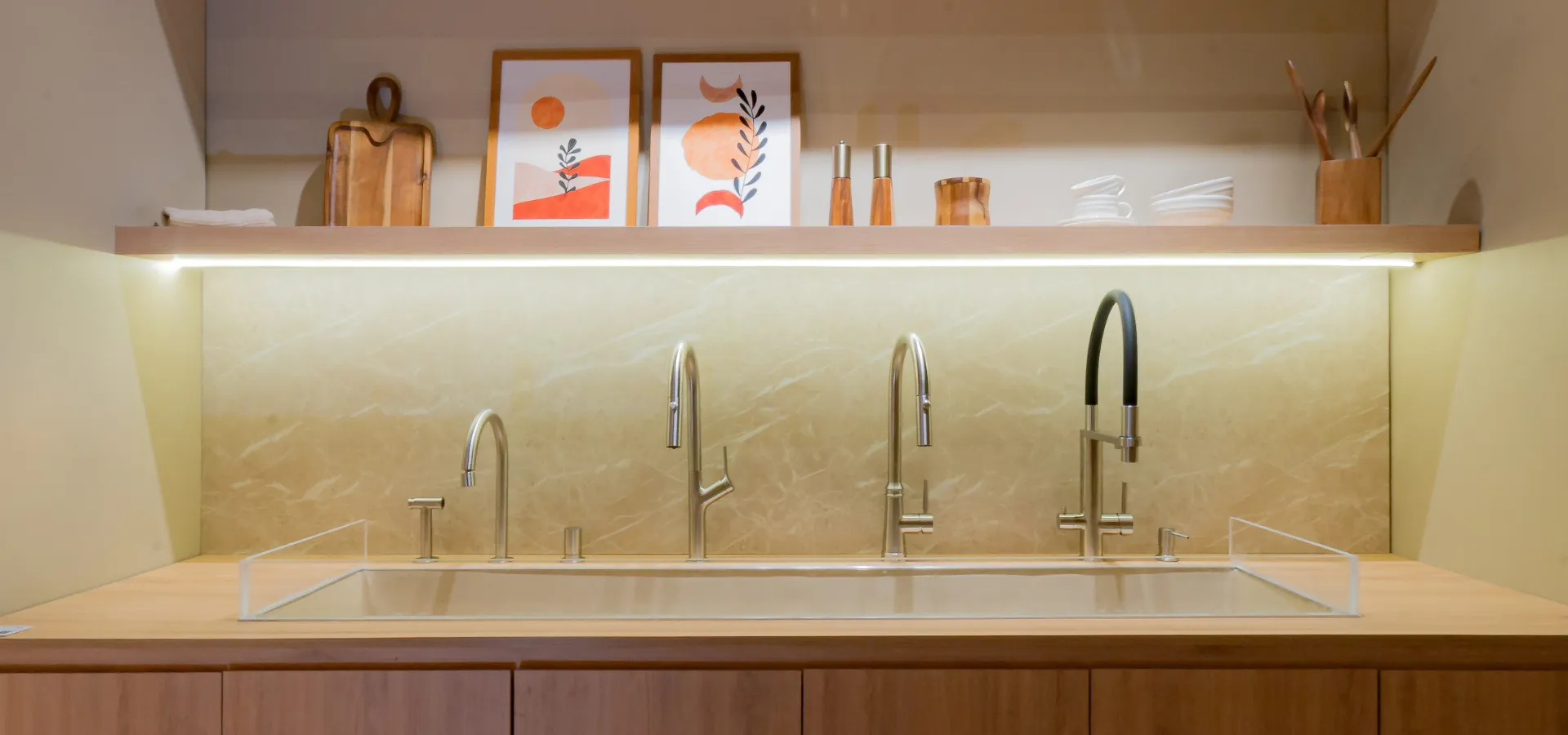 Cozinhas modernas Doka 2026: painel expositivo da coleção Doka Kitchen com quatro monocomandos em aço inox escovado sobre bancada de madeira clara com cuba retangular embutida — da esquerda para a direita, Monocomando Modu com bica direcionável e ducha manual, Monocomando Pull-Out Neora com mangueira retrátil, Monocomando Pull-Out Ferme com mangueira flexível preta e Monocomando para Filtro Aera com bica dupla; prateleira superior decorada com quadros, tábuas de madeira e utensílios, sob iluminação linear embutida.