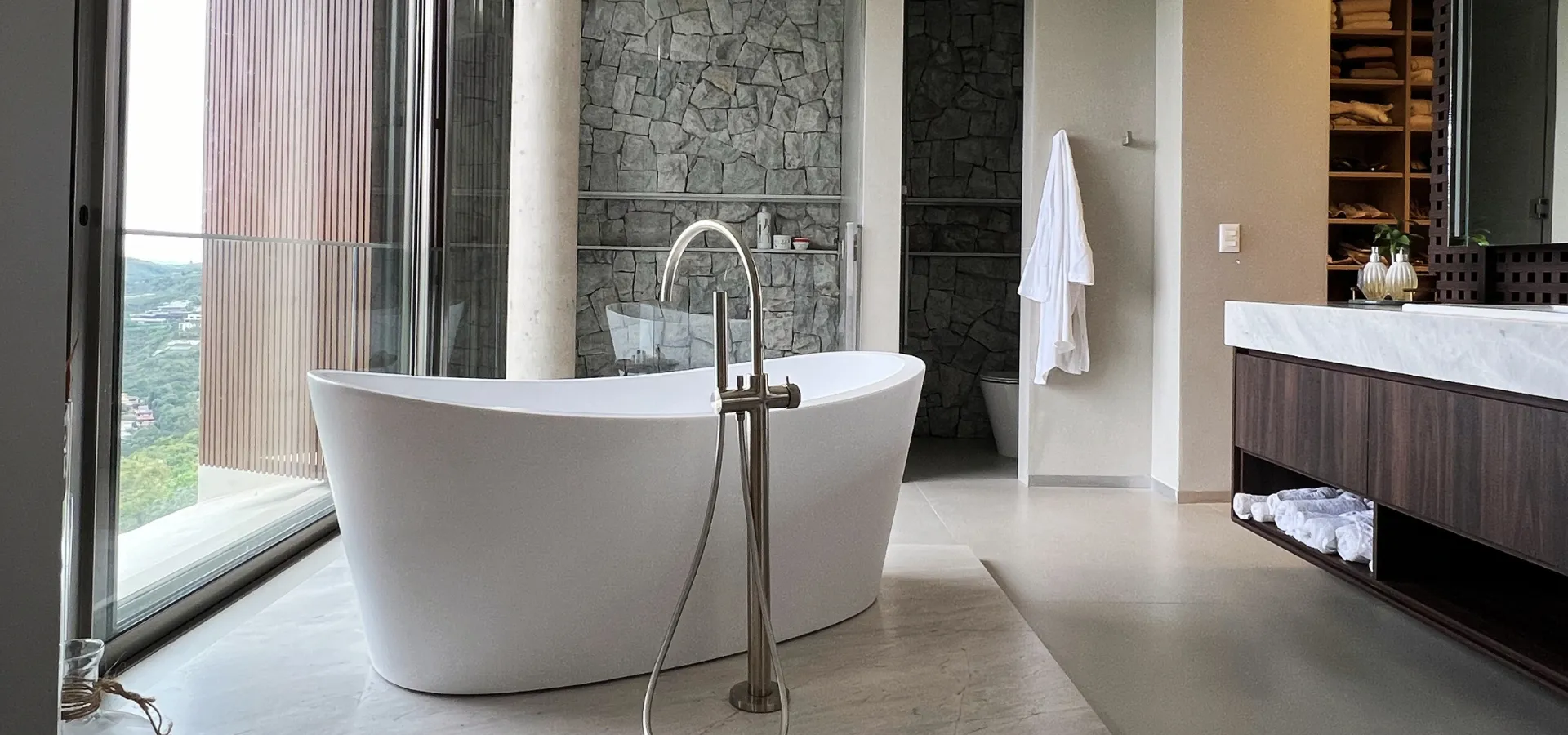 Banheira de freestanding Creta X na cor Matt White, com torneira de coluna em aço escovado, instalada em banheiro de alto padrão com piso em porcelanato claro, parede de pedra natural, ampla esquadria de vidro com vista para área verde e bancada suspensa em madeira escura com cuba em mármore.
