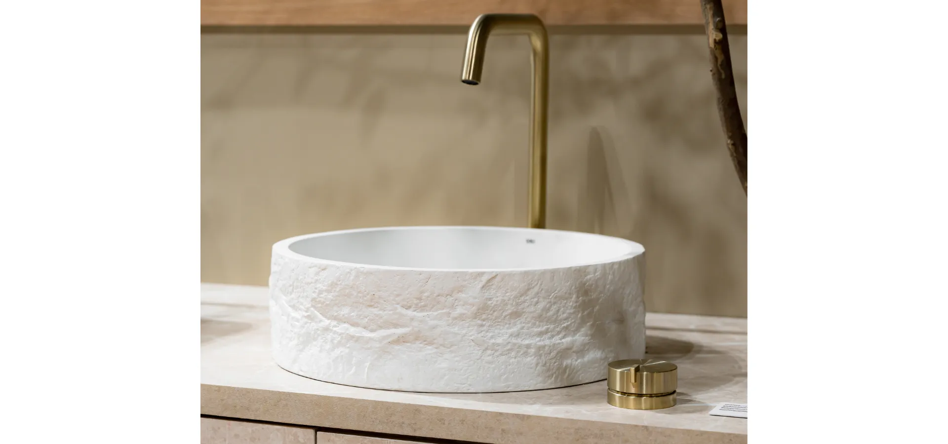 Cuba de apoio redonda Rock em acabamento branco com textura de pedra natural esculpida, sobre bancada em porcelanato claro, acompanhada de torneira de parede e válvula de escoamento Quantum em Brushed Gold, com fundo em parede de cimento queimado e detalhe em madeira natural. Saiba quais são as dimensões ideais para um banheiro confortável