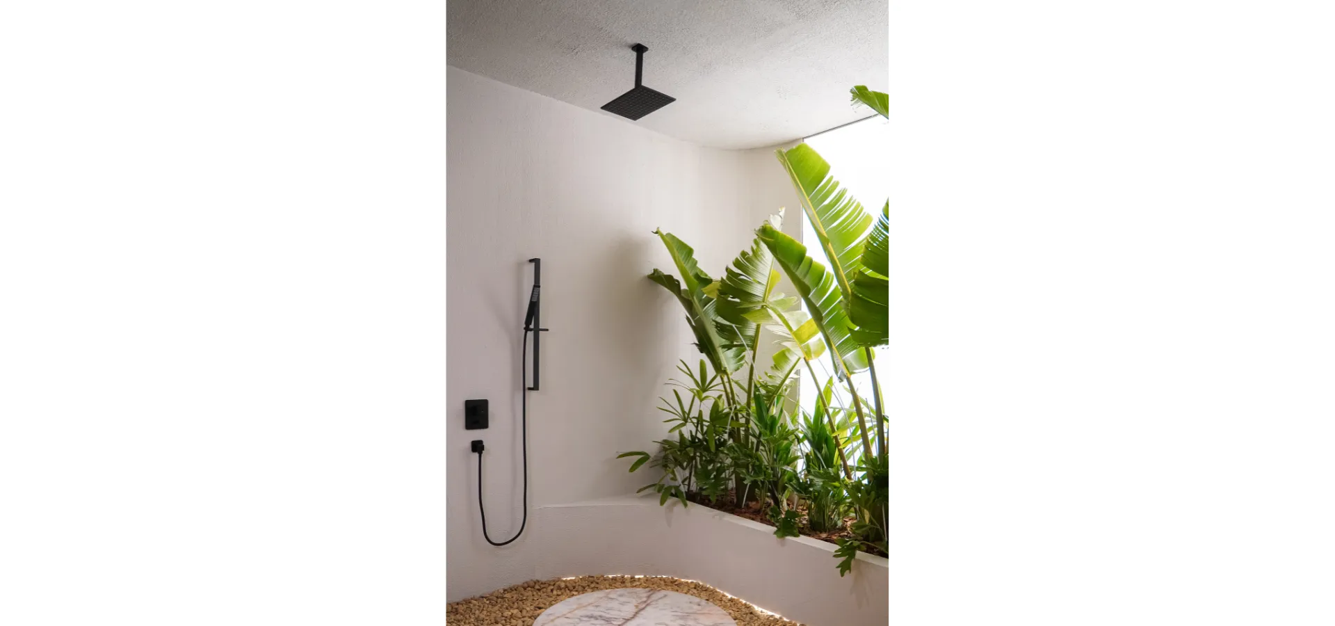 Banheiro contemporâneo com metais Rivvo em acabamento preto fosco, incluindo chuveiro de teto quadrado e ducha manual. O ambiente possui paredes claras, jardim interno com plantas tropicais e piso com pedras decorativas, criando uma atmosfera de spa integrada à natureza. A composição destaca soluções para um banheiro confortável, funcional e visualmente amplo.