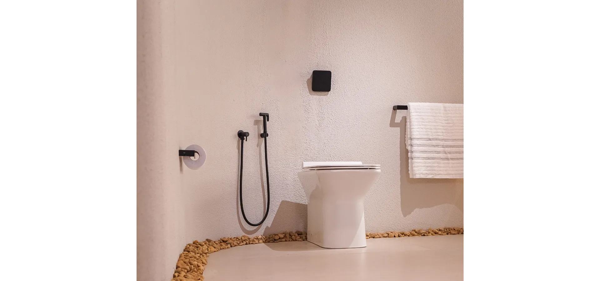Banheiro minimalista com metais Rivvo em preto fosco, incluindo ducha higiênica e acessórios coordenados. O ambiente apresenta vaso sanitário branco, paredes texturizadas em tom claro, toalheiro com toalha branca e detalhes em pedras decorativas no piso, reforçando a proposta de conforto, circulação adequada e design contemporâneo para banheiros compactos.