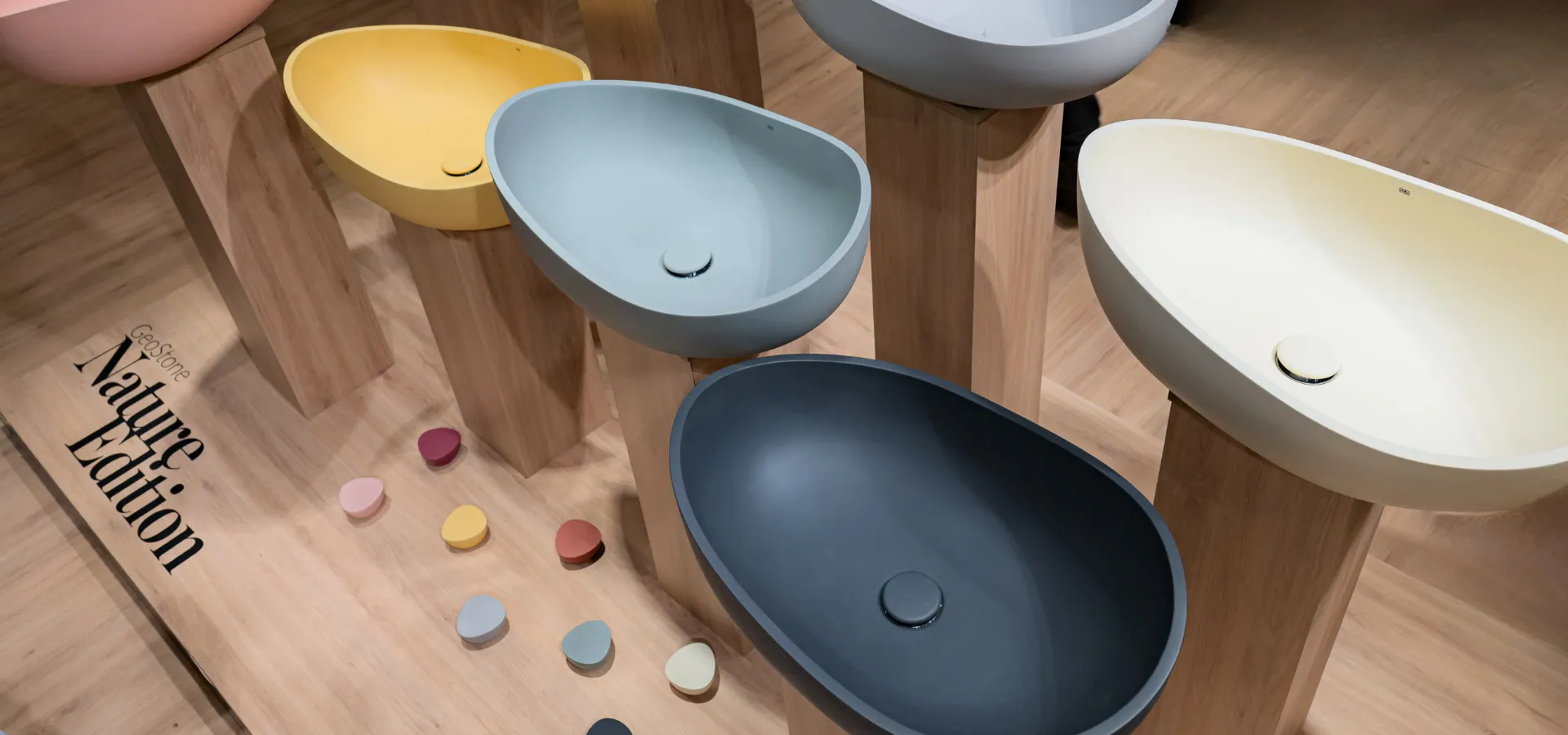 Banheiros modernos Doka 2026: exposição da coleção GeoStone Nature Edition com cubas de apoio orgânicas em acabamento fosco, apresentadas em cinco cores — rosa, amarelo, azul acinzentado, grafite e off-white — sobre suportes de madeira clara, acompanhadas de cabides orgânicos arredondados espalhados no piso nas mesmas cores da coleção.