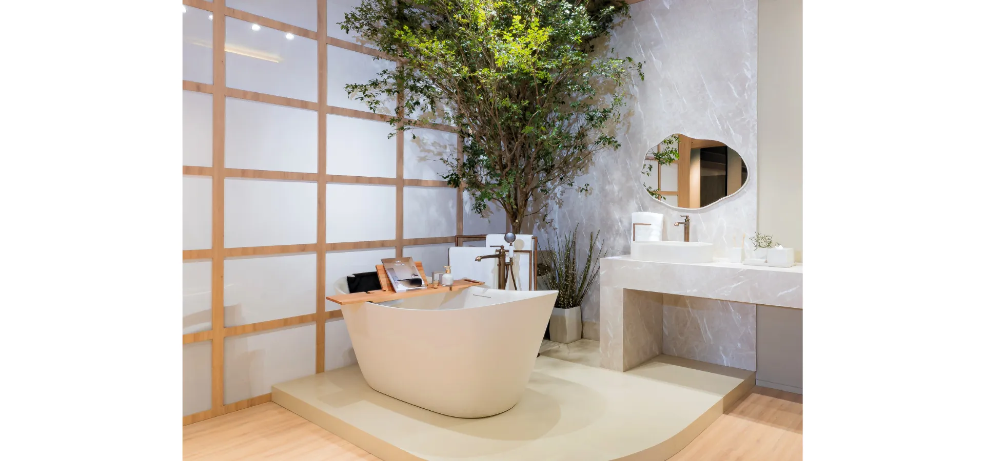 Banheira de imersão freestanding Doka linha Texture em Off-White em ambiente de showroom com bancada em mármore, cuba de apoio oval Off-White Terrazzo e metais Ever em ORB. Composição com árvore natural, estrutura ripada em madeira clara, revestimento mineral e piso em madeira. Todos os elementos do ambiente em coerência de materiais e acabamentos. Doka Collection Texture.